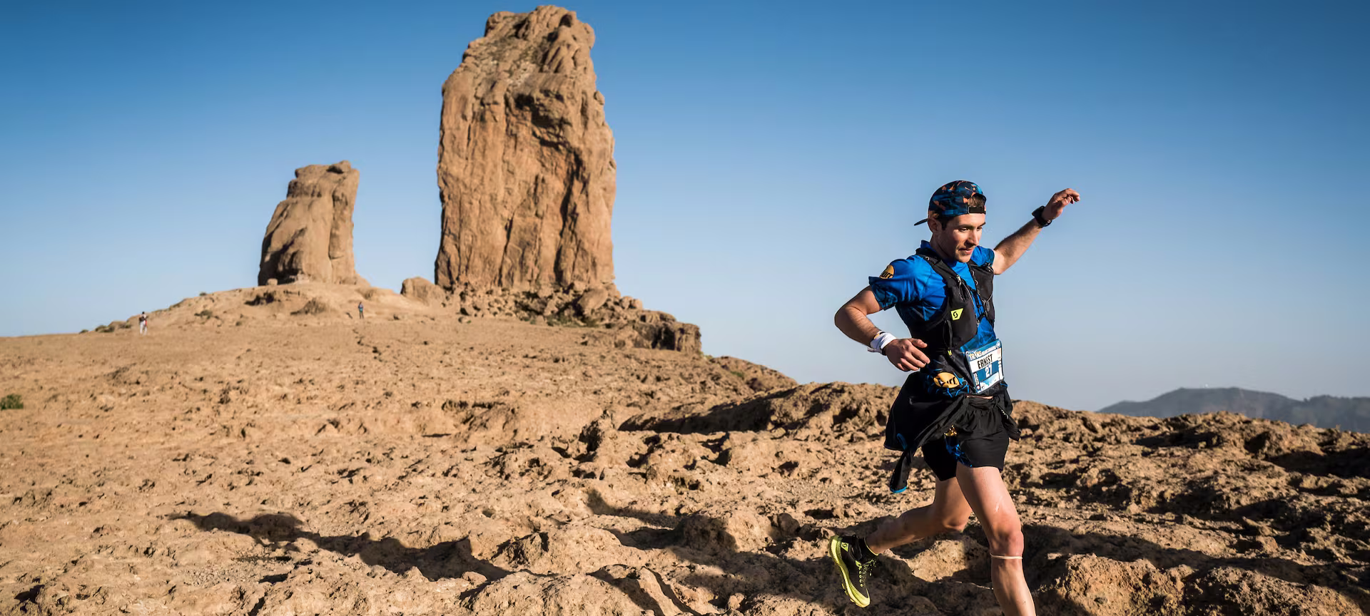 Un corredor corriendo sonriente por un sendero árido de la Transgrancanaria con una gran roca de fondo