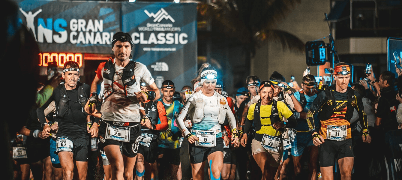 Un corredor corriendo sonriente por un sendero árido de la Transgrancanaria con una gran roca de fondo